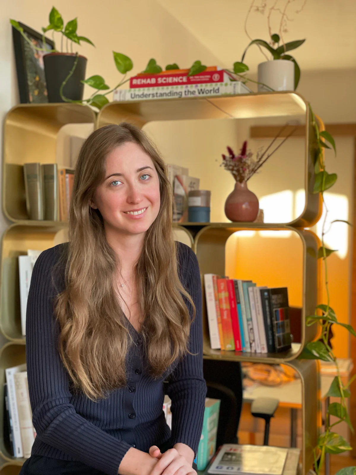 Anna Sierocinska in front of a gold bookshelf with plants and books, smiling warmly at the camera, her hands clasped.